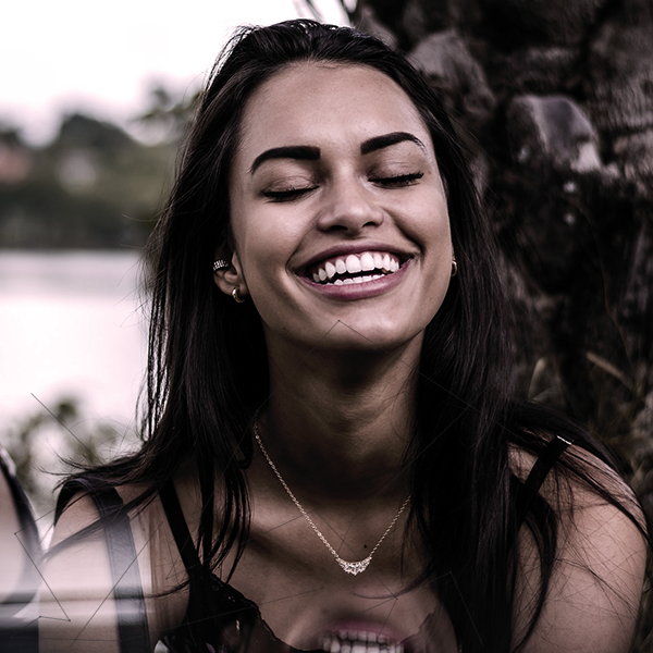 Foto de uma mulher sorrindo, ela está com os olhos fechados e demonstra uma alegria singular. Ao fundo, vemos uma linda paisagem verde desfocada. 