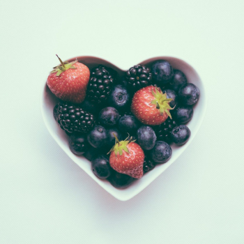 Vasilha branca em formato de coração com frutas dentro, como morando, amoras e blueberries em um fundo verde claro.